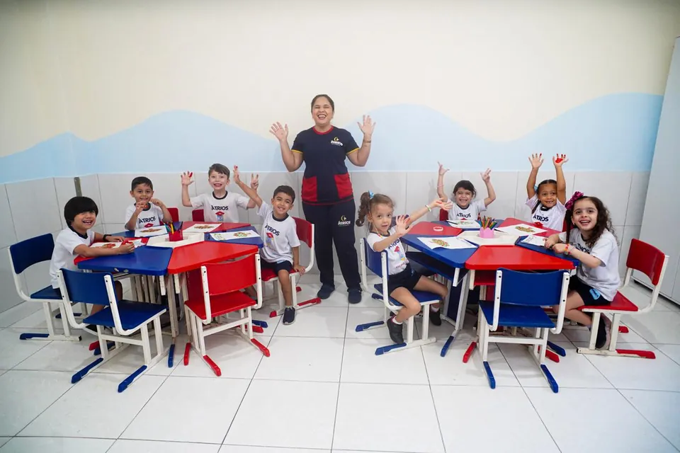 Sala de aula com mesas coloridas, professora e alunos acenando
